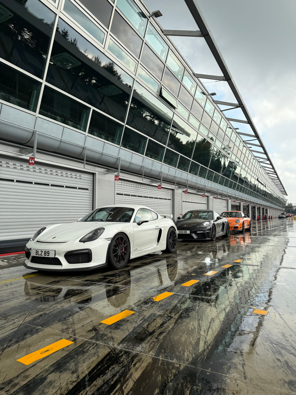Sports cars, including a white Porsche, parked at a wet UK racetrack pit lane, Eastwood Engineering